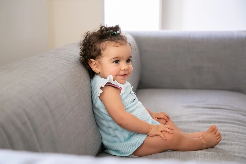 joyful-dark-curly-haired-baby-girl-wearing-pale-blue-cloth-sitting-grey-couch-home-looking-away-smiling-kid-home-childhood-concept